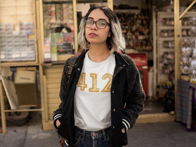 May include: A woman wearing a white t-shirt with the number "412" printed in gold on the front. She is also wearing a black jacket and blue jeans.