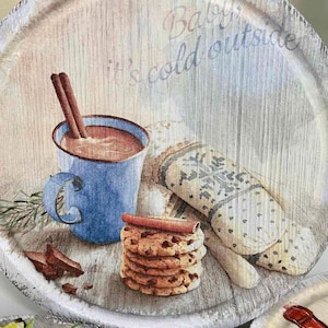 May include: A round wooden coaster with a white painted edge. The coaster features a painted image of a mug of hot chocolate with cinnamon sticks, a stack of chocolate chip cookies, and a mitten. The text "Baby, it's cold outside" is written in cursive on the coaster.