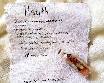 May include: A handwritten note on a piece of paper with the word "Health" at the top. The note lists various crystals and their properties, including pink salt, ginger, aventurine, clear quartz, rose, and a white candle. The note also includes a protection symbol and the text "mars sign". A small glass bottle filled with crystals and herbs is sitting on the paper. The note says "Keep in home or on person to promote healing and health."