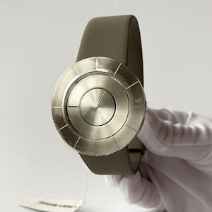 May include: A silver-toned wristwatch with a circular face and a gray leather band. The watch face has a unique design with radial lines and a central dial. The band is a muted gray color. The watch is held up by a gloved hand.