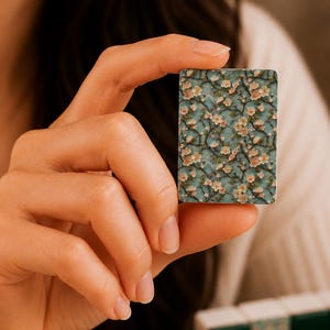 May include: A rectangular mahjong tile featuring a detailed floral pattern with light pink blossoms and brown branches set against a teal blue background. The tile has a smooth, flat surface and is held upright to display the intricate botanical design.
