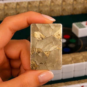 May include: A rectangular mahjong tile featuring a cracked, mosaic-like pattern in shades of beige, gray, and metallic gold. The surface has a smooth, polished finish with distinct angular segments that create a complex, multi-toned appearance.