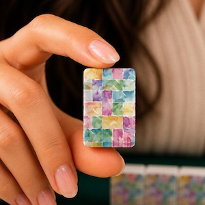 May include: A rectangular mahjong tile featuring a colorful mosaic pattern of small, interlocking squares in shades of pink, yellow, blue, and green. The tile is held upright, displaying a vibrant, multi-colored design on its front surface.