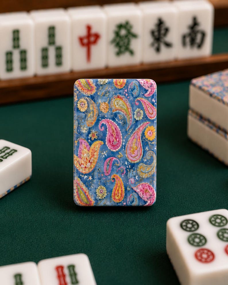 May include: A rectangular mahjong tile featuring a vibrant blue background decorated with a colorful paisley pattern in shades of pink, yellow, and orange. The tile is positioned upright among other standard white mahjong tiles on a green surface.