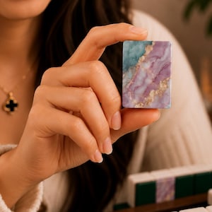K&ouml;nnte beinhalten: Ein rechteckiger Mahjong-Spielstein mit einem Marmormuster in T&uuml;rkis- und Violettt&ouml;nen sowie goldfarbenen Aderungen. Der Spielstein hat eine glatte, gl&auml;nzende Oberfl&auml;che und wird aufrecht gehalten, um das komplexe, mehrfarbige Design zu zeigen.