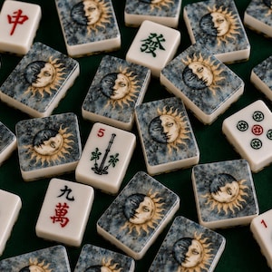 May include: A collection of rectangular mahjong tiles scattered across a surface. Many tiles feature a celestial sun and moon face illustration with a blue-grey textured background. Other tiles display traditional Chinese characters and symbols.