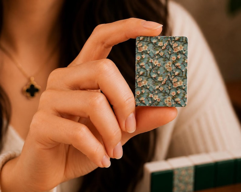 May include: A rectangular mahjong tile featuring a detailed floral pattern with light pink blossoms and brown branches set against a teal blue background. The tile has a smooth, flat surface and is held upright to display the intricate botanical design.