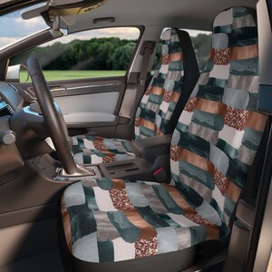 May include: Car seat covers with a watercolor-style design. The covers feature a pattern of rectangular shapes in shades of teal, brown, gray, and a shimmering gold. The car interior includes a steering wheel, dashboard, and a view of the outside.