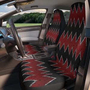 May include: Car seat covers with a black and red zigzag pattern. The covers are installed in a car, with the steering wheel and dashboard visible. The design features a repeating zigzag pattern in red and black.