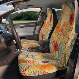 Puede incluir: Fundas de asiento de coche con un vibrante estampado floral. El diseño presenta grandes flores en tonos amarillos, rojos y blancos, sobre un fondo amarillo. Las fundas son visibles dentro de un coche, con el volante y el salpicadero a la vista.