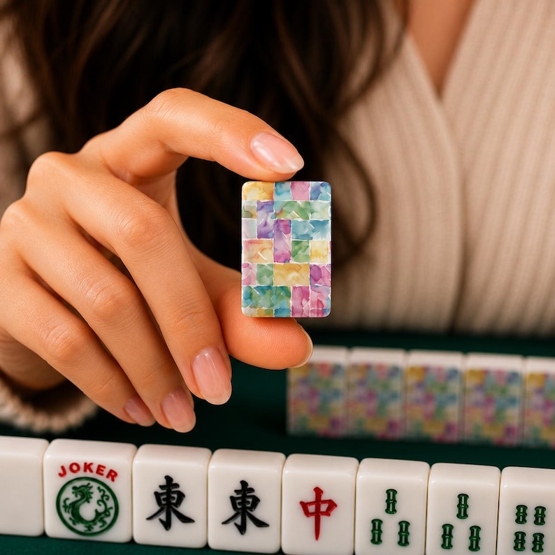 May include: A rectangular mahjong tile featuring a colorful mosaic pattern of small, interlocking squares in shades of pink, yellow, blue, and green. The tile is held upright, displaying a vibrant, multi-colored design on its front surface.