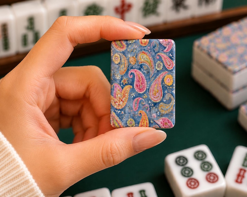 May include: A rectangular mahjong tile featuring a vibrant blue background with a detailed, multi-colored paisley pattern in shades of pink, yellow, orange, and green. The tile has a glossy finish and is held upright to display the intricate design.