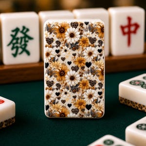 May include: A rectangular phone case featuring a floral design with yellow and white daisies, leopard print hearts, and black hearts. The case is surrounded by white Mahjong tiles with green and red Chinese characters.