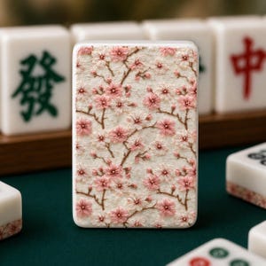K&ouml;nnte beinhalten: Nahaufnahme einer wei&szlig;en Mahjong-Kachel mit floralem Design. Die Kachel hat eine strukturierte Oberfl&auml;che mit einem Muster aus rosa Kirschbl&uuml;ten und braunen Zweigen. Weitere Kacheln mit chinesischen Schriftzeichen sind im Hintergrund zu sehen.