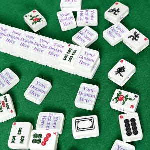 Puede incluir: Una colecci&oacute;n de fichas de mahjong rectangulares blancas esparcidas sobre una superficie verde. Varias fichas presentan el texto morado Your Design Here, mientras que otras muestran s&iacute;mbolos tradicionales en negro, rojo y verde.