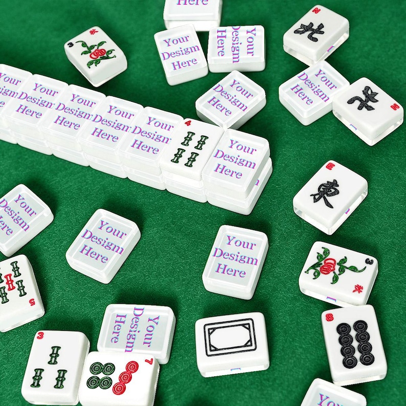 Puede incluir: Una colecci&oacute;n de fichas de mahjong rectangulares blancas esparcidas sobre una superficie verde. Varias fichas presentan el texto morado Your Design Here, mientras que otras muestran s&iacute;mbolos tradicionales en negro, rojo y verde.