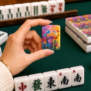 May include: A rectangular mahjong tile featuring a vibrant, multi-colored floral painting with shades of blue, purple, yellow, and red. The tile is held upright, displaying a detailed impressionistic flower design against a dark green surface.