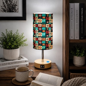May include: A decorative table lamp with a colorful geometric pattern on the shade. The lamp has a wooden base with a USB port and a silver metal stem. The shade features squares and circles in red, teal, orange, and black.