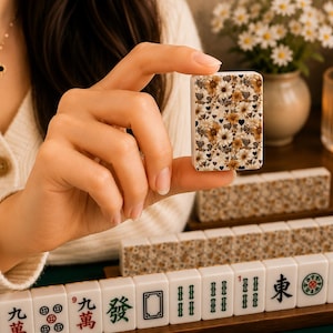 May include: A single mahjong tile held between fingers, featuring a floral pattern with daisies and black hearts on a cream background. Other tiles are visible, along with a wooden rack and a vase of flowers.