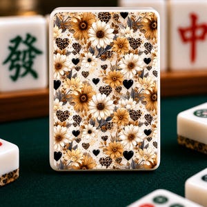 May include: A rectangular Mahjong tile with a floral design featuring sunflowers, white daisies, and black heart accents. The tile has a cream-colored background with leopard print details. Other Mahjong tiles are visible in the background.