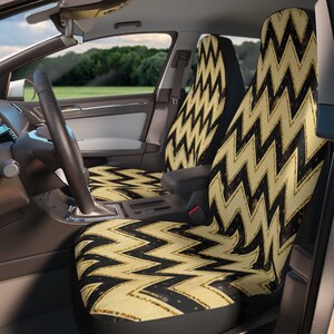 Puede incluir: Fundas de asiento de coche con un patrón de chevrón negro y dorado. Las fundas están instaladas en un coche, con el volante y el salpicadero visibles. El diseño es un zigzag repetitivo, creando un aspecto geométrico audaz.