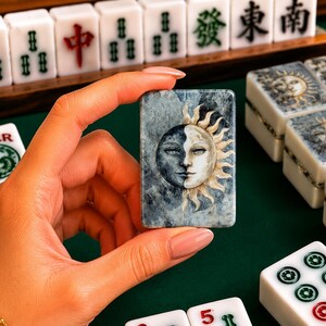 May include: A rectangular mahjong tile featuring a dual-toned sun and moon face illustration with a marbled blue and gray background. The tile is held upright, displaying a detailed celestial design with one side dark and the other side light.