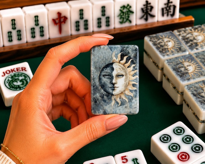 May include: A rectangular mahjong tile featuring a dual-toned sun and moon face illustration with a marbled blue and gray background. The tile is held upright, displaying a detailed celestial design with one side dark and the other side light.