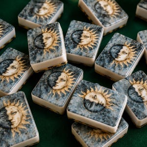 May include: A collection of rectangular mahjong tiles featuring a printed design of a sun and moon face on a marbled blue and grey background. Each tile has a white base and a smooth, glossy surface, displaying a celestial motif with golden rays.