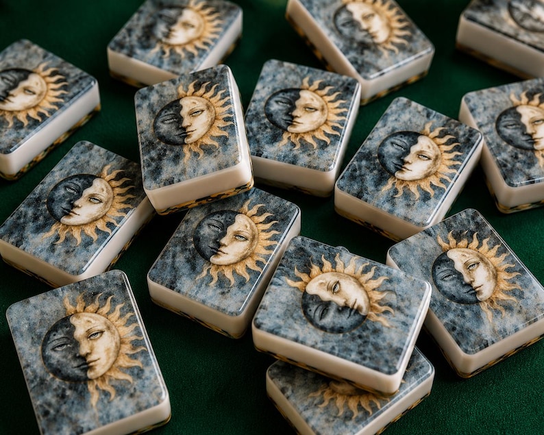 May include: A collection of rectangular mahjong tiles featuring a printed design of a sun and moon face on a marbled blue and grey background. Each tile has a white base and a smooth, glossy surface, displaying a celestial motif with golden rays.