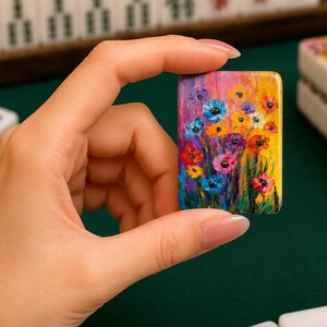 May include: A rectangular mahjong tile featuring a vibrant, multi-colored floral painting. The artwork displays a dense cluster of stylized flowers in shades of blue, yellow, red, purple, and pink against a bright yellow and green background.