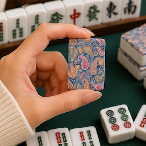 May include: A rectangular mahjong tile featuring a detailed blue, pink, and yellow paisley pattern on its face. The tile has a smooth, glossy finish and is held upright, displaying the intricate floral-inspired design against a dark green surface.