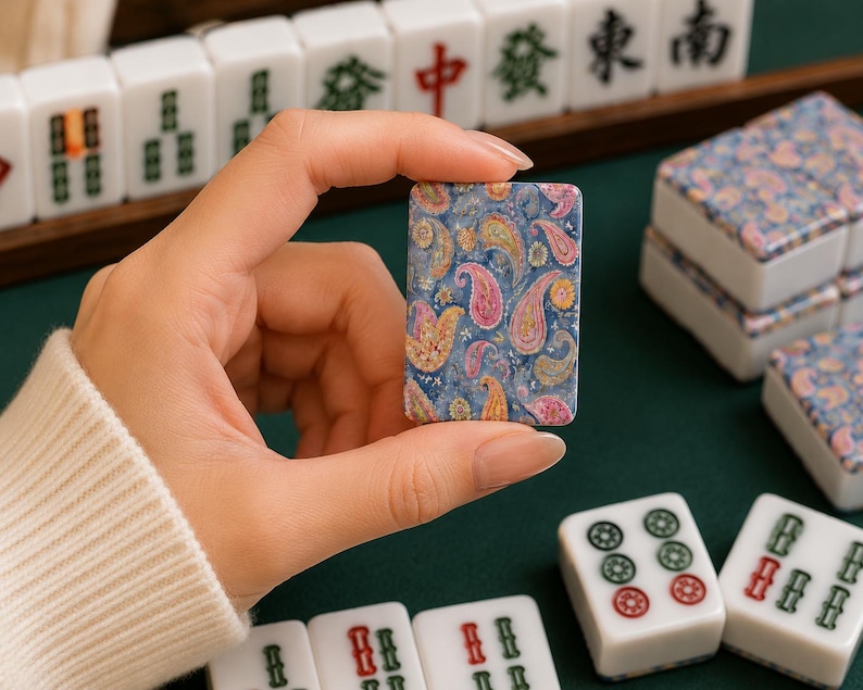 May include: A rectangular mahjong tile featuring a detailed blue, pink, and yellow paisley pattern on its face. The tile has a smooth, glossy finish and is held upright, displaying the intricate floral-inspired design against a dark green surface.