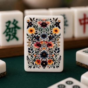Op de afbeelding: Een close-up van een witte Mahjong-tegel met een bloemenontwerp. De tegel heeft een levendige opstelling van kleurrijke bloemen in tinten rood, oranje, geel en blauw, tegen een achtergrond van groene bladeren. Andere tegels zijn zichtbaar op de achtergrond.