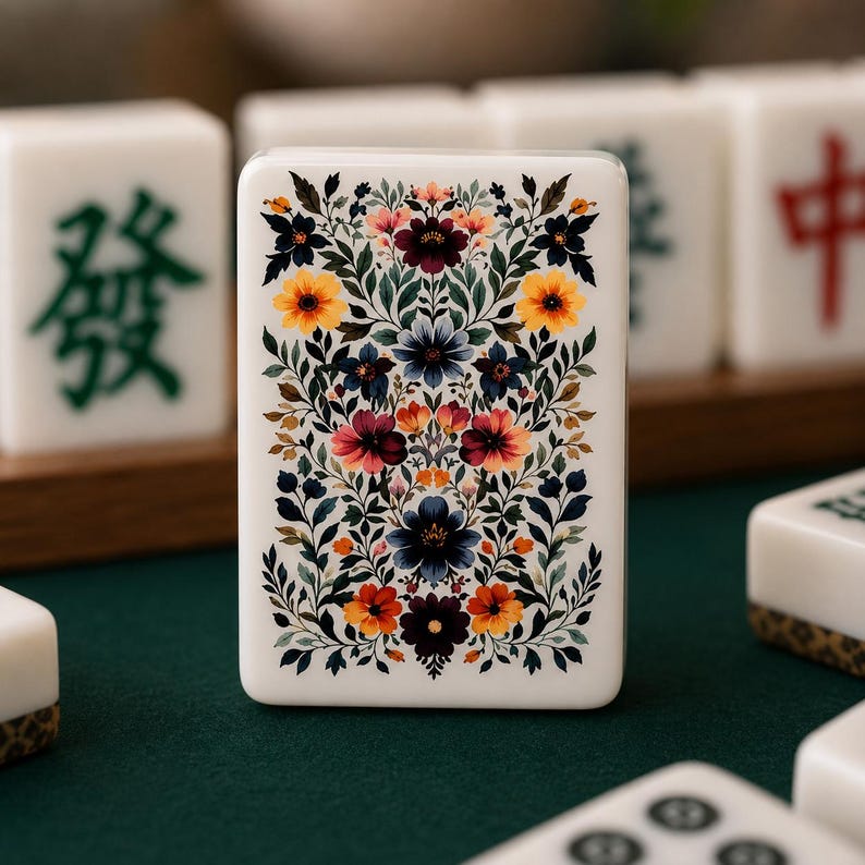 Op de afbeelding: Een close-up van een witte Mahjong-tegel met een bloemenontwerp. De tegel heeft een levendige opstelling van kleurrijke bloemen in tinten rood, oranje, geel en blauw, tegen een achtergrond van groene bladeren. Andere tegels zijn zichtbaar op de achtergrond.
