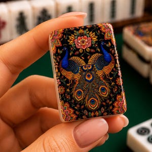 Puede incluir: Una ficha de mahjong rectangular que presenta una ilustraci&oacute;n detallada y colorida de dos pavos reales con plumaje azul y dorado vibrante, rodeados de intrincados patrones florales sobre un fondo negro, con base blanca y bordes redondeados.