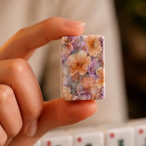 Op de afbeelding: Een rechthoekige mahjongtegel met een gedetailleerd bloemenpatroon van perzikkleurige, lichtpaarse en witte bloesems op een gedempte blauwe achtergrond. De tegel heeft een gladde, glanzende afwerking en afgeronde hoeken met een dichte botanische print.