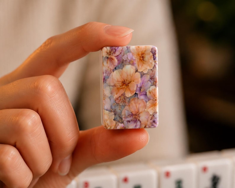 Op de afbeelding: Een rechthoekige mahjongtegel met een gedetailleerd bloemenpatroon van perzikkleurige, lichtpaarse en witte bloesems op een gedempte blauwe achtergrond. De tegel heeft een gladde, glanzende afwerking en afgeronde hoeken met een dichte botanische print.