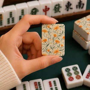 May include: A rectangular mahjong tile featuring a cream-colored background with a detailed botanical print of orange flowers and green leaves. The tile is held upright, displaying the floral pattern clearly against other standard white mahjong tiles.