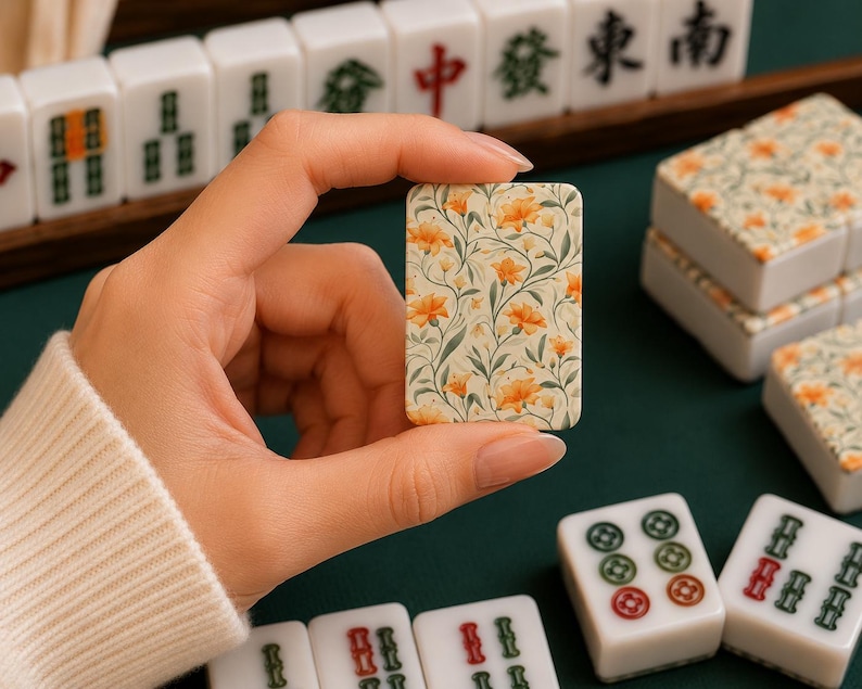 May include: A rectangular mahjong tile featuring a cream-colored background with a detailed botanical print of orange flowers and green leaves. The tile is held upright, displaying the floral pattern clearly against other standard white mahjong tiles.