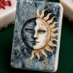 May include: A rectangular mahjong tile featuring a detailed illustration of a sun and moon face merged together. The background has a mottled blue and gray marble-like texture, while the sun rays are rendered in shades of cream and gold.