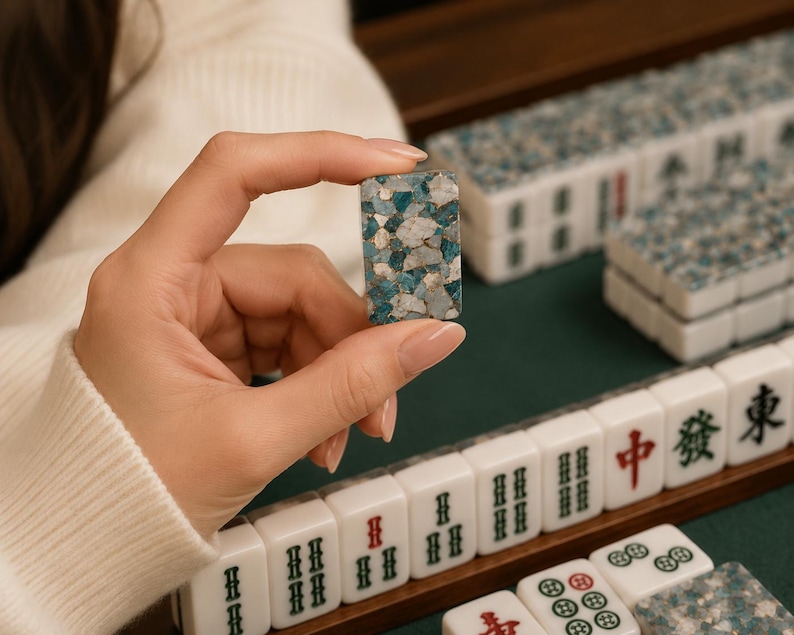 May include: A rectangular mahjong tile featuring a mosaic pattern of blue, white, and grey stone fragments. The tile is held upright, displaying its textured surface, while other standard white mahjong tiles with black and red characters are arranged in rows.