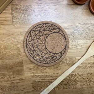 May include: A round cork coaster with a crescent moon and mandala design etched in black. The coaster is on a wooden surface with a wooden spoon to the side. The coaster is approximately 10 cm in diameter.
