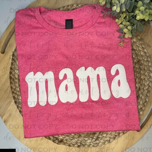 Op de afbeelding: Roze T-shirt met een witte "mama" grafische afbeelding in een retro fontstijl.