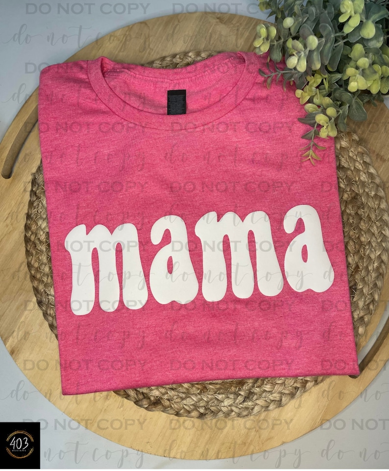 Op de afbeelding: Roze T-shirt met een witte "mama" grafische afbeelding in een retro fontstijl.