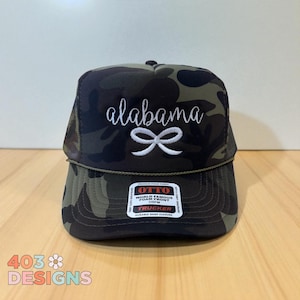 May include: A camouflage trucker hat with the word "alabama" in white script and a white bow graphic. The hat has a mesh back and a curved brim. The hat has a tag that says "OTTO" and "TRUCKER".