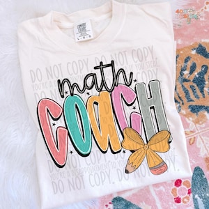 May include: Cream-colored t-shirt with the words "math COACH" in a colorful, playful design. The design includes a pencil and bow graphic. The shirt also has the text "DO NOT COPY" repeated throughout.