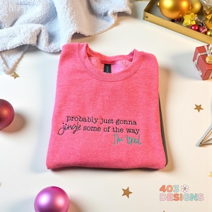 Op de afbeelding: Een roze crewneck sweatshirt met de tekst "probably just gonna jingle some of the way, I'm tired" in zwarte en blauwgroene letters. De sweatshirt is opgevouwen en ligt op een wit oppervlak met kerstversieringen.