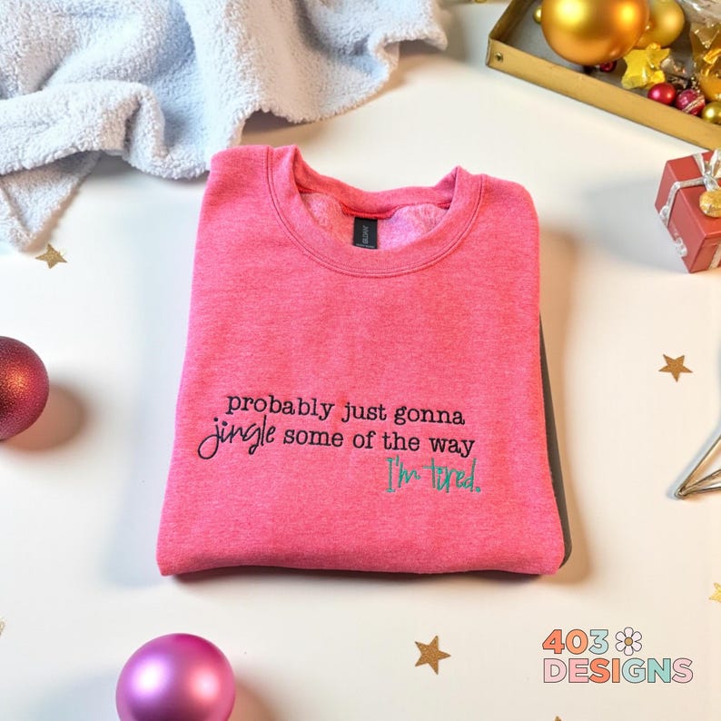 Op de afbeelding: Een roze crewneck sweatshirt met de tekst "probably just gonna jingle some of the way, I'm tired" in zwarte en blauwgroene letters. De sweatshirt is opgevouwen en ligt op een wit oppervlak met kerstversieringen.