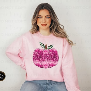 Puede incluir: Una sudadera rosa con un diseño de calabaza rosa brillante. La calabaza tiene un tallo y hojas verdes.