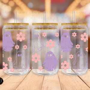 May include: Three clear glass beverage cups with bamboo lids and straws. Each cup features a purple ghost and pink flower design. The cups are arranged on a wooden surface.
